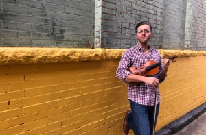 A man in a plaid shirt leans against a yellow brick wall while holding a violin and bow.