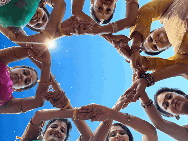 A group of women in colorful traditional attire stand in a circle, joining hands and looking down towards the camera against a clear blue sky.