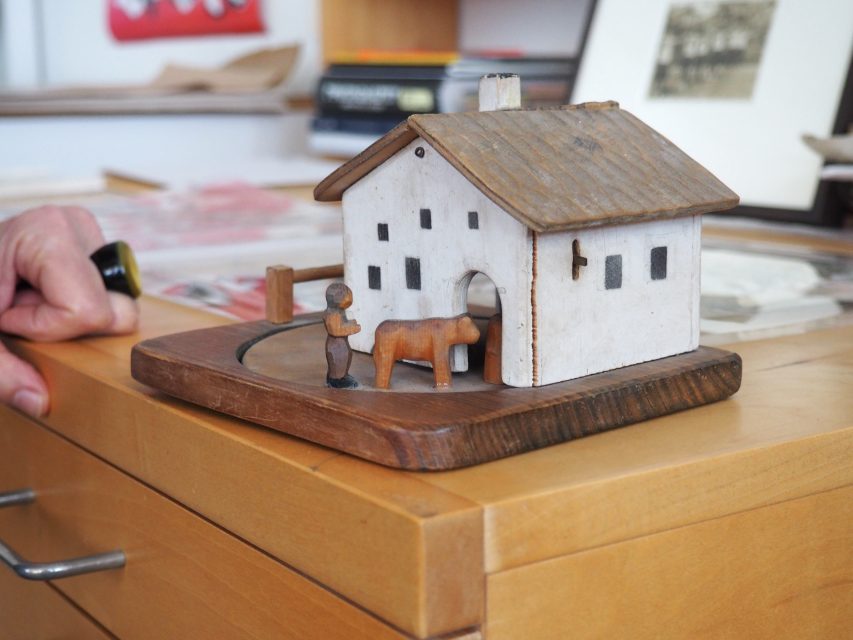 A small wooden model of a house with a figure and a cow on a wooden base sits on a wooden cabinet, with a hand and papers visible in the background.