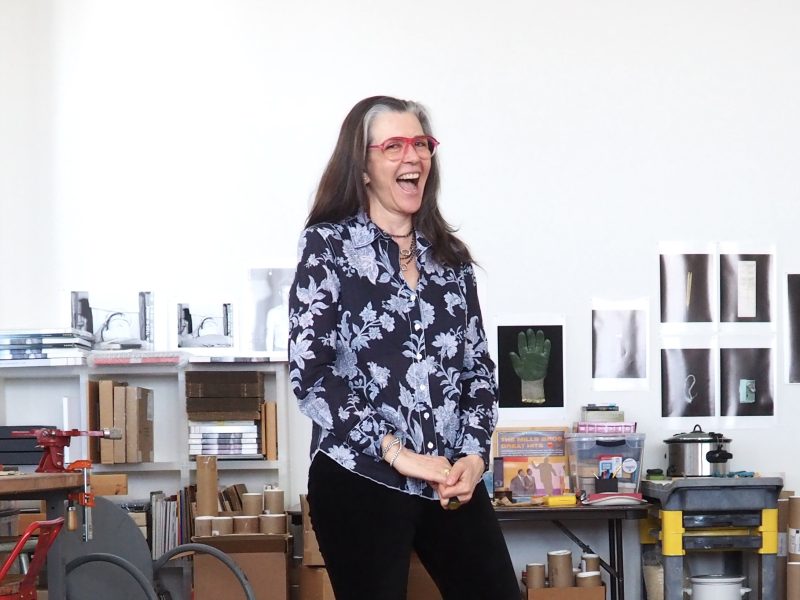 A woman with long dark hair and red glasses, wearing a floral shirt, stands smiling in a workspace with art supplies, books, and photos on the wall behind her.