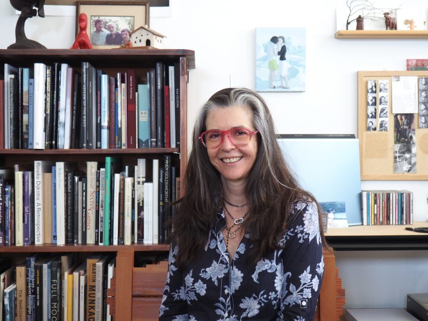 A woman with long gray hair and red glasses sits smiling in front of bookshelves and a bulletin board in a well-lit room.