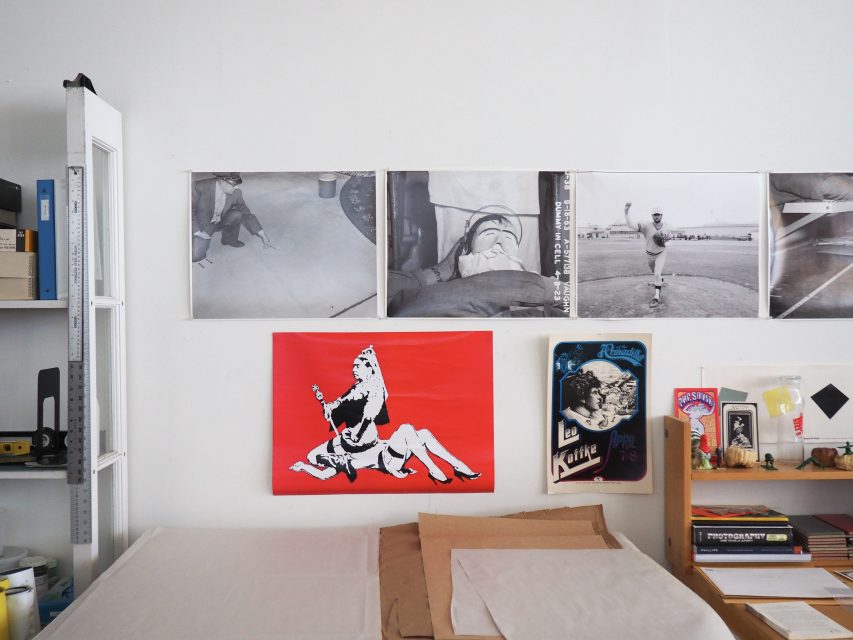 A workspace with art supplies, books, and paper beneath a wall displaying three black-and-white photos and a red poster with a graphic illustration.