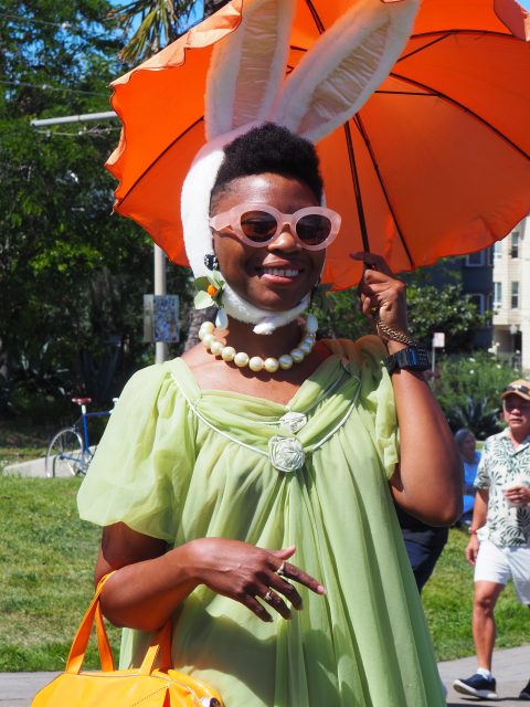 Person wearing a light green dress, large white bunny ears, and sunglasses holds an orange umbrella outdoors on a sunny day.