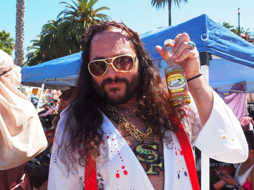 A man with long hair, sunglasses, and a fake crown of thorns holds up a bottle of pepper plant hot sauce at an outdoor event, with a tent and palm trees in the background.