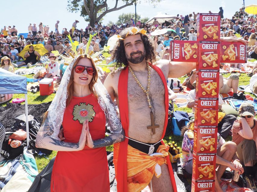 Two people in costume pose at an outdoor event; one is dressed as a saint, the other as Jesus holding a tall stack of Cheez-It boxes, with a large crowd sitting on the grass behind them.