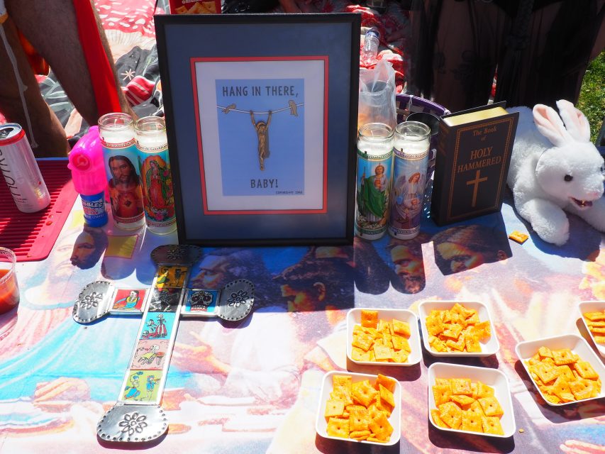 A table displays religious candles, a framed "Hang in there, baby!" print, a decorative cross, a Bible, a plush white rabbit, and trays of Cheez-It snacks.