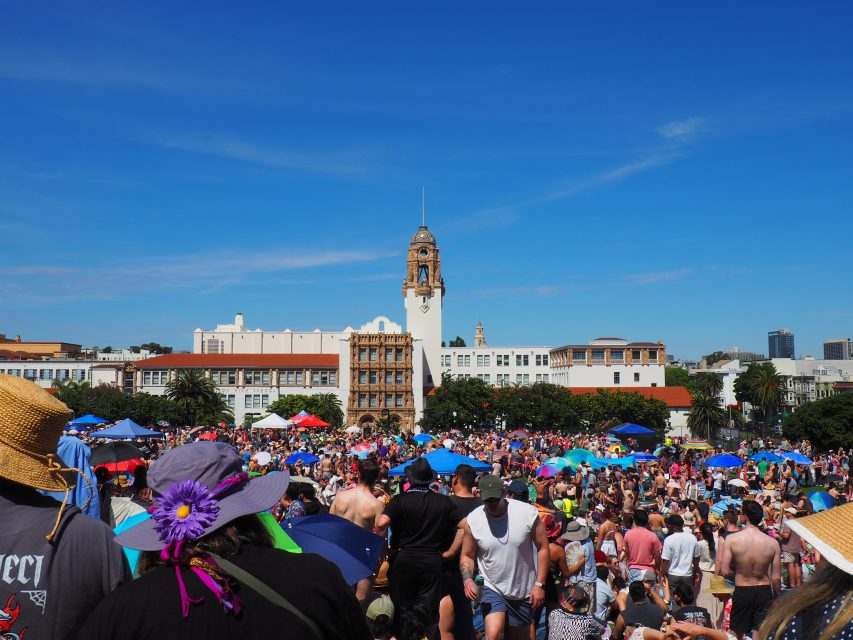Large crowd of people gathered outdoors under blue skies, many with hats and umbrellas, in front of a historic building with a clock tower.