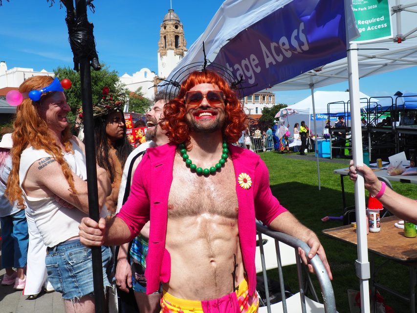 Person with red wig, pink jacket, green necklace, and flower pin smiles at an outdoor event with others in festive costumes. Stage and tents visible in background.