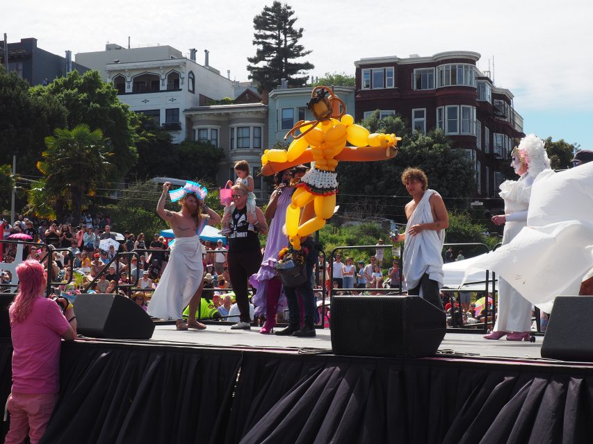 Several people in colorful costumes, including a large yellow balloon outfit, perform on an outdoor stage with a crowd and residential buildings in the background.