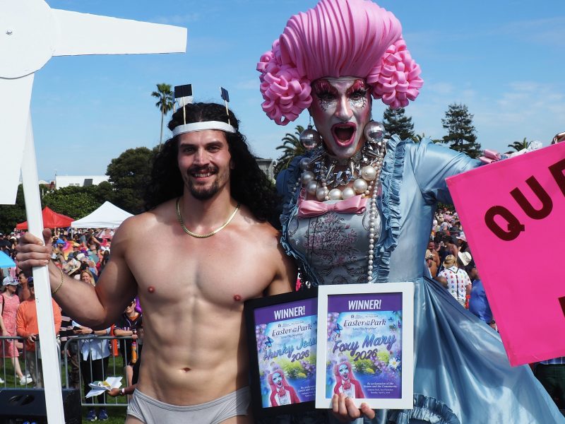 A shirtless man and a person in elaborate drag attire pose together at an outdoor event, each holding a "Foxy Mary 2023" winner plaque.