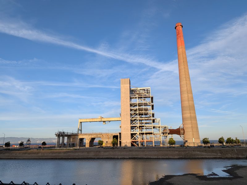 Industrial facility with a tall smokestack and exposed steel framework beside a body of water, under a clear blue sky.