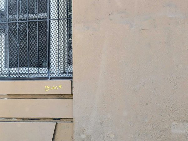 A beige wall with graffiti that says "BLACK" in yellow near a window covered by metal bars and mesh.