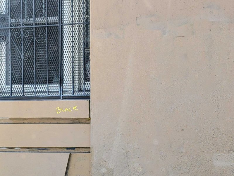 A beige wall with a small section labeled "BLACK" in yellow writing, next to a window with metal bars and mesh.