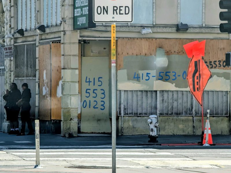 Two people stand on a sidewalk near a boarded-up building with phone numbers spray-painted on it; traffic signs and cones are visible in the foreground.