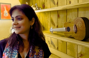 A woman with dark hair stands indoors near a wooden shelf displaying a ukulele; a framed picture of a ukulele hangs on the wall behind her.