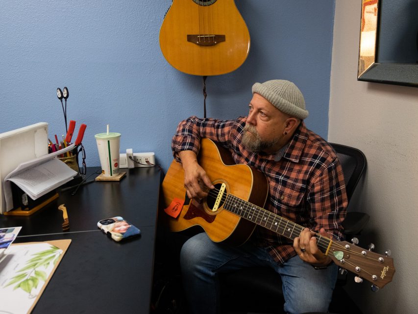 A man wearing a beanie and plaid shirt sits in an office playing an acoustic guitar, with papers, a drink, and a phone on the desk beside him.