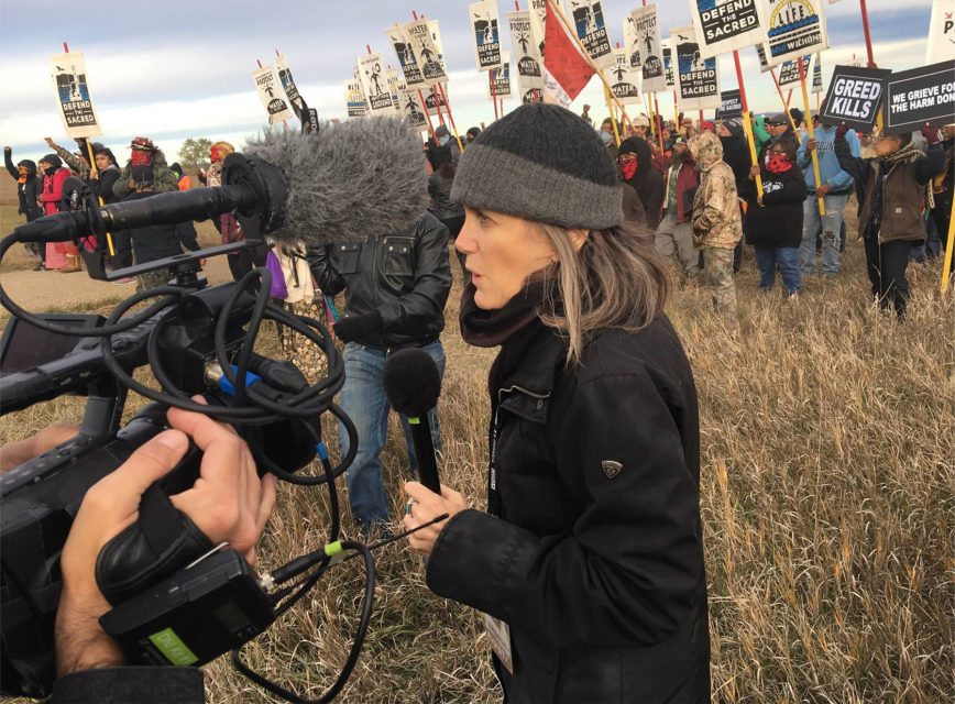 A reporter holds a microphone and speaks to a camera crew in a field while a group of protesters holding signs stands in the background.