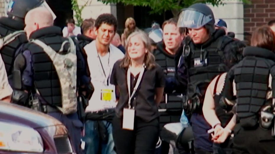 A woman with credentials around her neck is escorted by multiple police officers in riot gear during a crowded outdoor event.