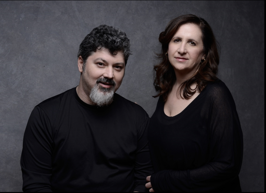 Two adults, a man with gray hair and a beard and a woman with long brown hair, pose together against a plain gray backdrop, both wearing black tops.
