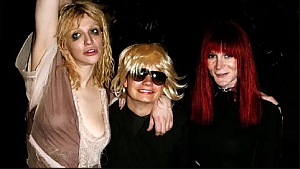 Three people posing for a photo; one with blonde messy hair and sheer dress, one with a short blonde wig and sunglasses, and one with long red hair and a black outfit.