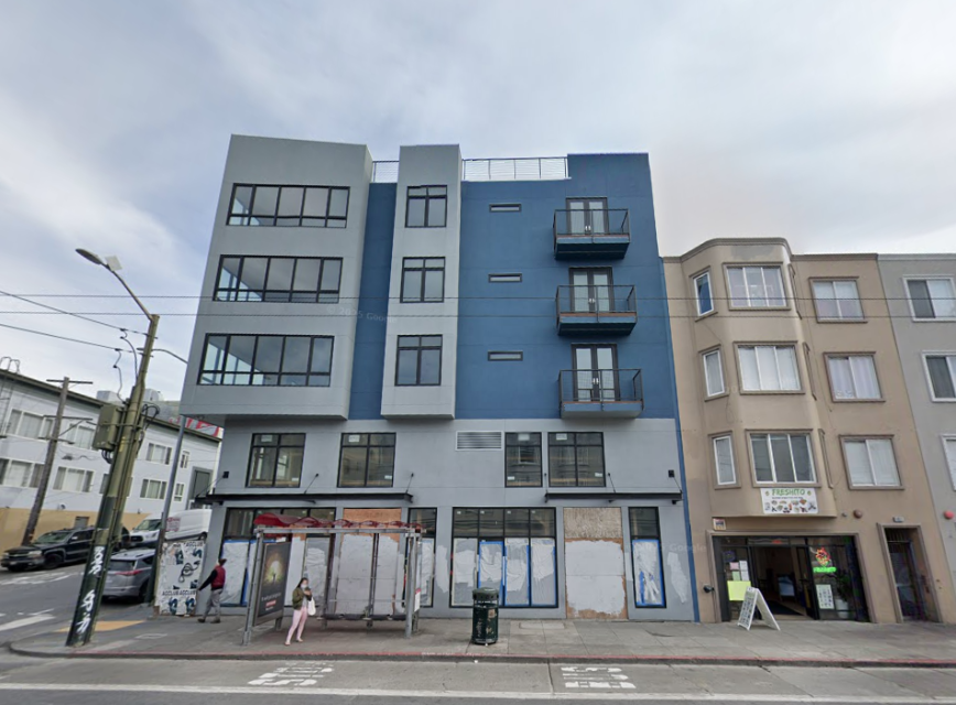 A four-story blue and gray apartment building with balconies stands next to older beige buildings; storefronts on the ground level are boarded up, and a bus stop is in front.