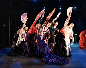 A group of flamenco dancers in colorful dresses perform on stage, holding fans above their heads and leaning to one side under stage lighting.