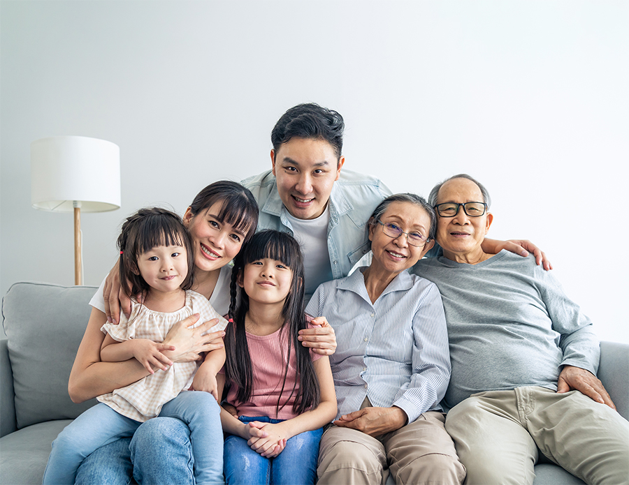 Three generations of an Asian family, including two children, parents, and grandparents, sit together on a couch at home, smiling at the camera.