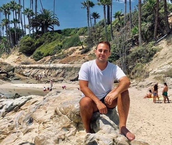 A man in a white t-shirt and shorts sits on rocks at a sandy beach with palm trees and people in the background under a clear sky.
