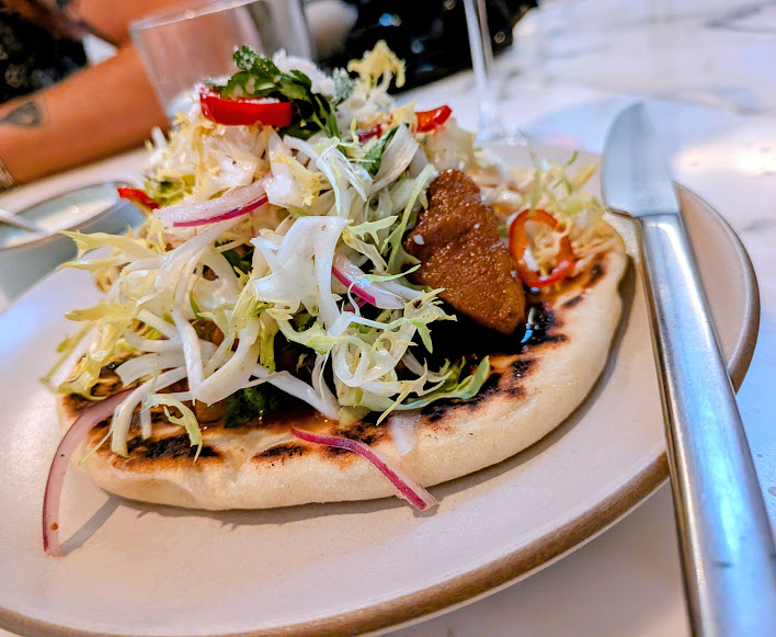 A flatbread topped with grilled meat, shredded lettuce, red onions, and sliced red chili, served on a plate with a knife and fork.