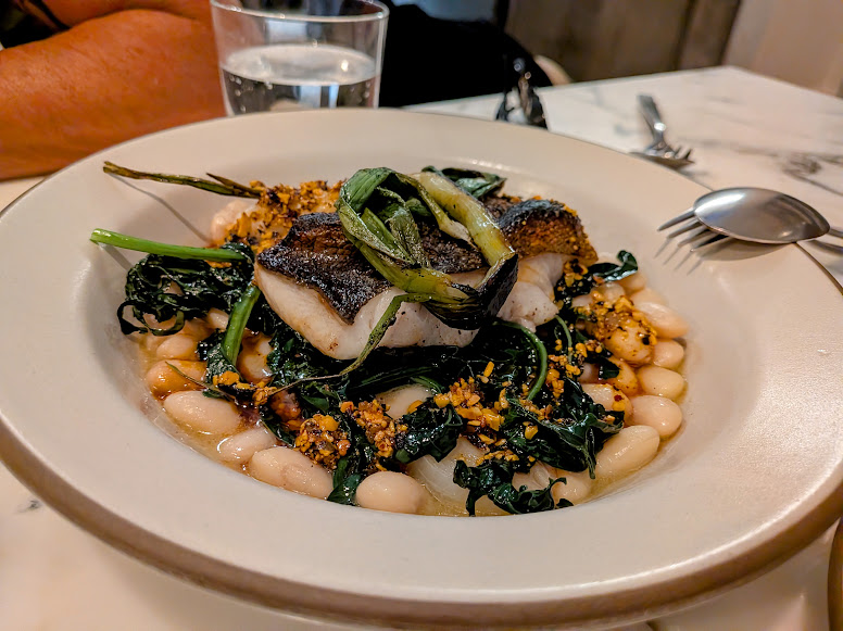 A plate of seared fish served on white beans and sautéed greens, garnished with charred scallions, with a glass of water and cutlery in the background.