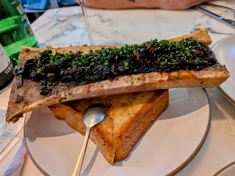 A plate with a large roasted bone marrow topped with herbs and chopped vegetables, served on toasted bread with a spoon.