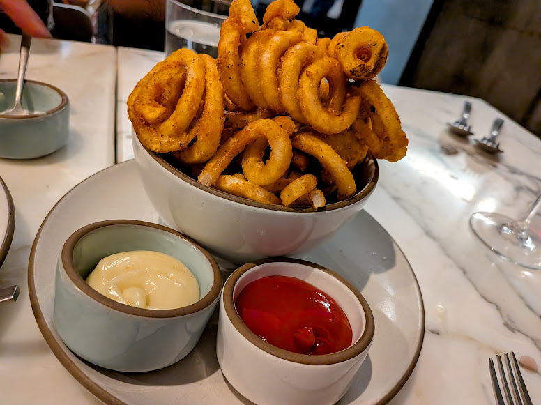A bowl of seasoned curly fries served with sides of mayonnaise and ketchup on a white plate, placed on a marble tabletop.