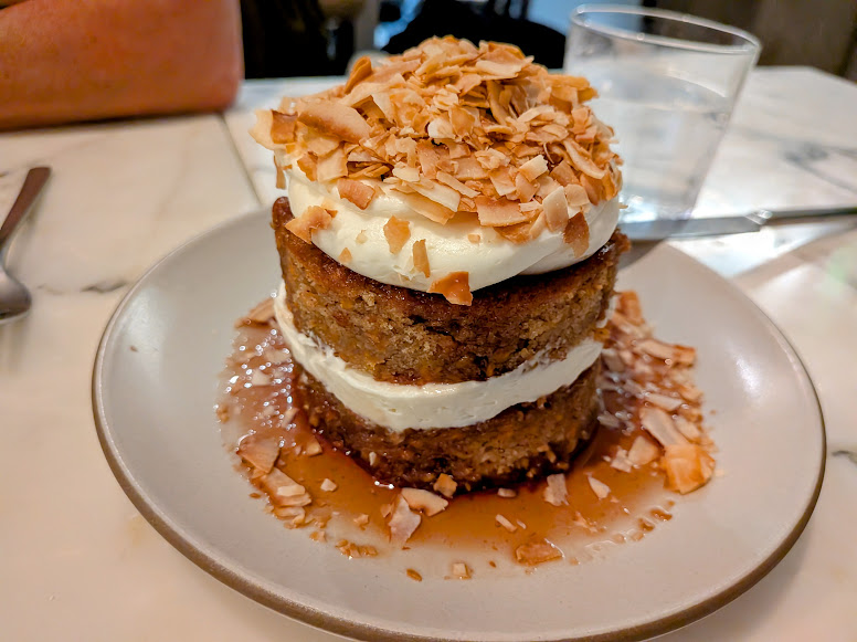 A layered cake with cream filling, whipped topping, and toasted coconut flakes, served on a plate with syrup, next to a glass of water on a marble table.