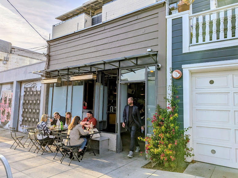 People sitting at outdoor café tables and talking while a man exits the building; plants and a garage are visible beside the café.