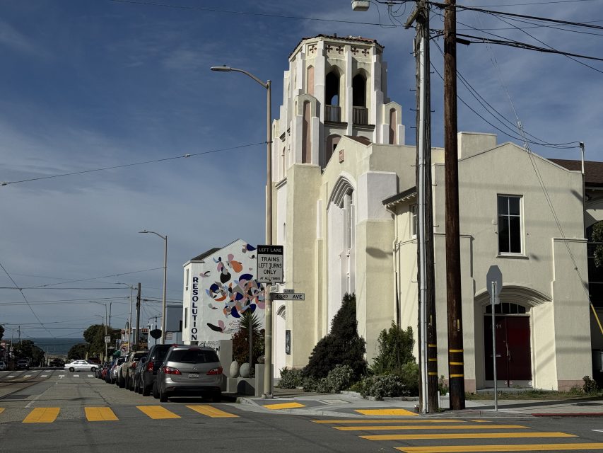 A beige church with a tall bell tower stands on a street corner next to parked cars and a crosswalk under a clear blue sky.