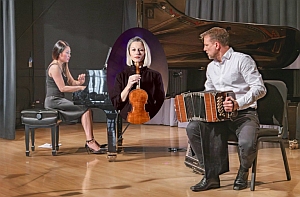 A woman plays piano, a man plays bandoneon, and another woman holding a violin is featured in an oval inset above them on stage.