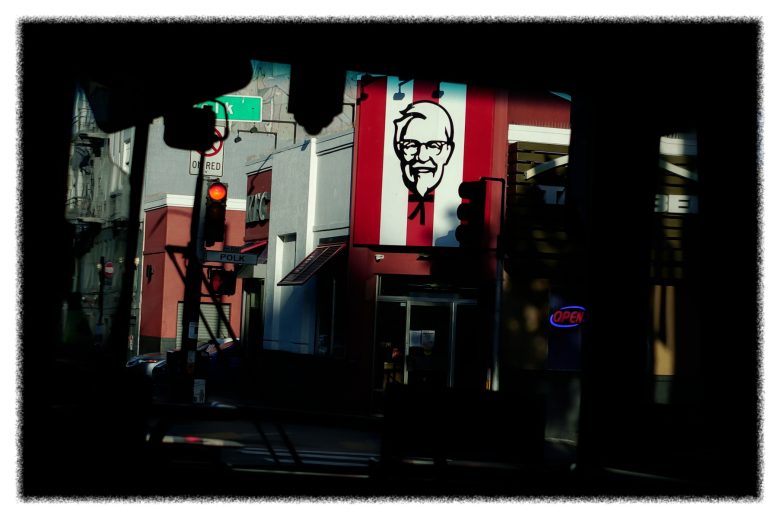 A KFC restaurant with a large Colonel Sanders sign is seen across the street; traffic light shows red and an "OPEN" sign is illuminated in the window.