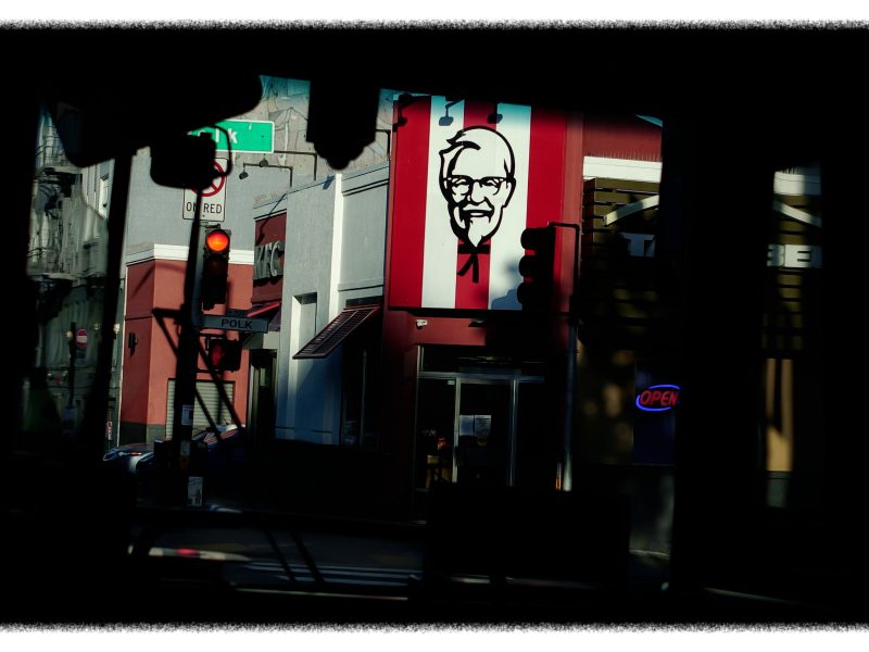 A KFC restaurant with a large Colonel Sanders sign is seen across the street; traffic light shows red and an "OPEN" sign is illuminated in the window.
