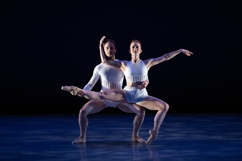 Two ballet dancers in white costumes perform a duet on stage, with the female dancer balanced on one leg and the male dancer supporting her.