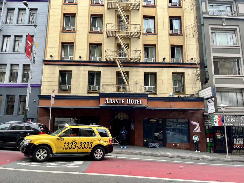 A yellow taxi drives past the entrance of Adante Hotel on a city street with mid-rise buildings and pedestrians nearby.