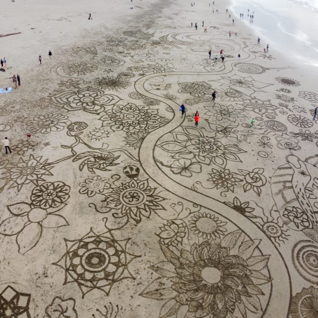 Aerial view of a beach with large, intricate floral and geometric patterns drawn in the sand; people walk among the designs near the shoreline.