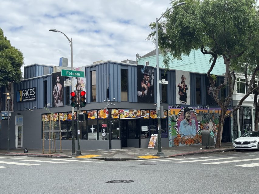 A corner building at an intersection displays murals, food photos, and portraits above the entrance, with a street sign reading "Folsom" and a traffic light in the foreground.