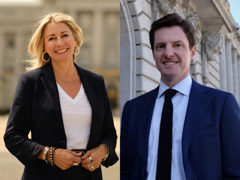 A woman and a man stand outdoors in front of stone buildings, both wearing business attire and smiling at the camera.