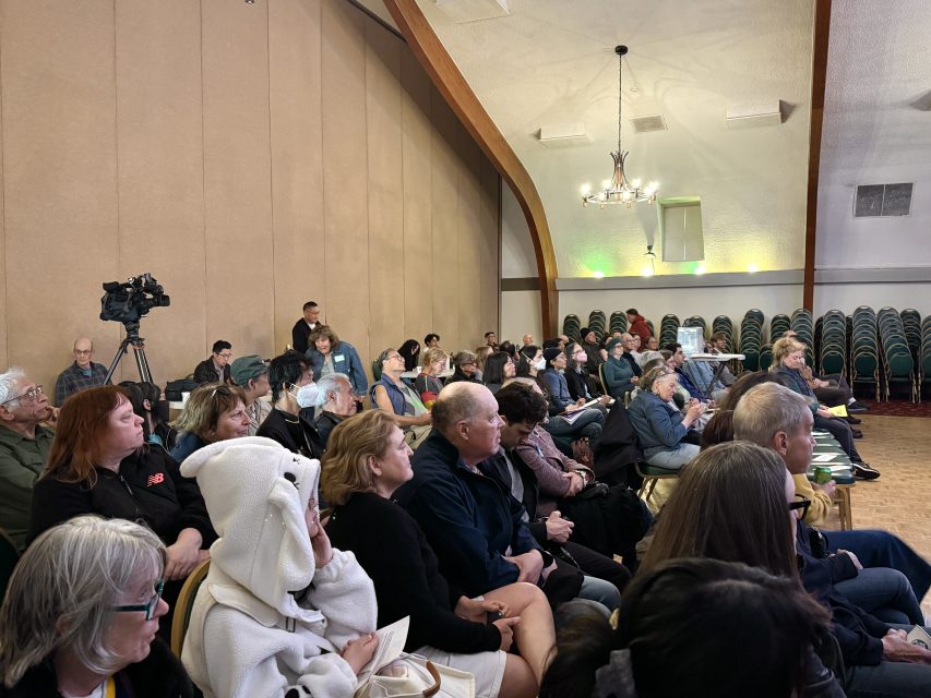 A group of people seated in rows inside a hall, attentively watching something out of frame; a camera on a tripod is set up in the back.