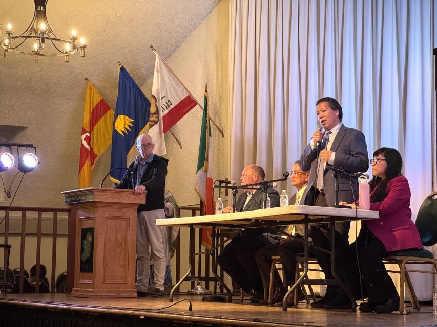 Five people participate in a panel discussion on stage; one man stands at a podium, while four others sit at a table with microphones. Flags are displayed in the background.