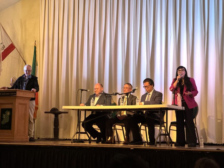 Five people participate in a panel discussion on stage; four are seated at a table with microphones, while one stands and speaks into a microphone. California and Mexican flags are visible.