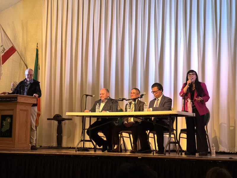 Five people participate in a panel discussion on stage; four are seated at a table with microphones, while one stands and speaks into a microphone. California and Mexican flags are visible.