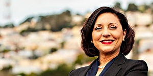 A woman in a dark blazer stands outdoors, smiling with her arms crossed, with a blurred cityscape in the background.