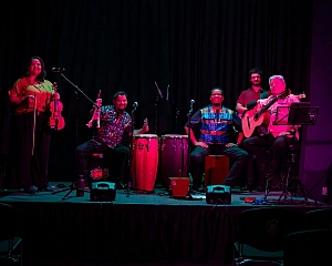 Five musicians perform on a dimly lit stage with various instruments, including a violin, congas, guitar, and flute. The background is dark, and the group is facing the audience.