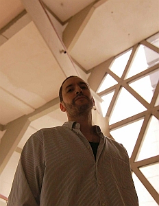 A man in a striped shirt stands indoors, viewed from a low angle, with geometric windows and ceiling beams visible in the background.